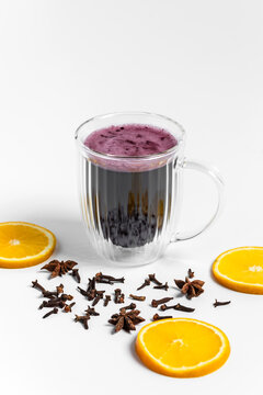 Red hot mulled wine for a cozy cold evening to warm up in a transparent double wall cup or mug on white background, star anise and cloves spices around, fresh citrus orange wheels 