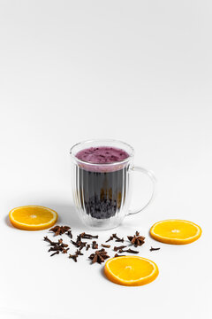 Red hot mulled wine for a cozy cold evening to warm up in a transparent double wall cup or mug on white background, star anise and cloves spices around, fresh citrus orange wheels 