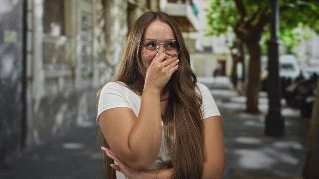 Woman covers mouth with hand on street wearing glasses and white t shirt; playful embarrassment shyness.