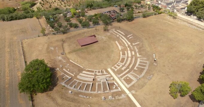 Aerial view of the semicircular remains of an ancient amphitheater. The stone foundations and seating rows are carved into the dry earth. It's the archaeological park of Venosa, in Basilicata, Italy.