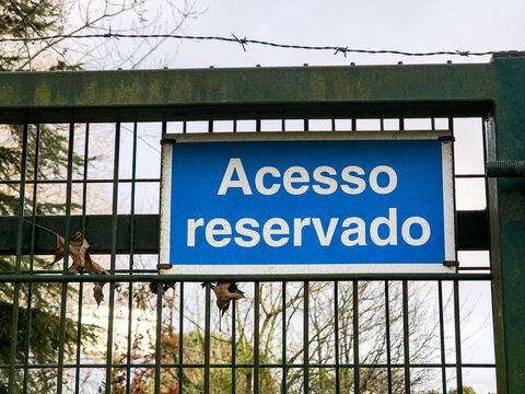 Blue sign reading &ldquo;Acesso reservado&rdquo; mounted on a metal fence with barbed wire, outdoor restricted area warning in Portuguese indicating private access and controlled entry to the enclosed space.