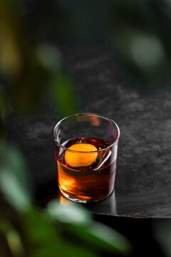 Classic old fashioned alcoholic cocktail in a transparent rocks glass with a big ice cube and orange peel garnish, lifestyle image on black wooden table in a bar 