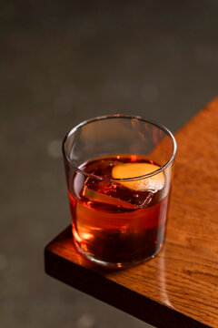 Old fashioned alcoholic cocktail in a transparent rocks glass with a big ice cube and orange peel garnish, lifestyle image on wooden table