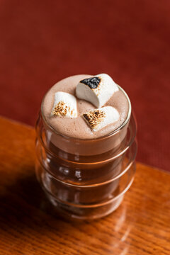 Hot cocoa chocolate drink in a transparent double wall glass with toasted marshmallows on top as a garnish, lifestyle image on wooden table 