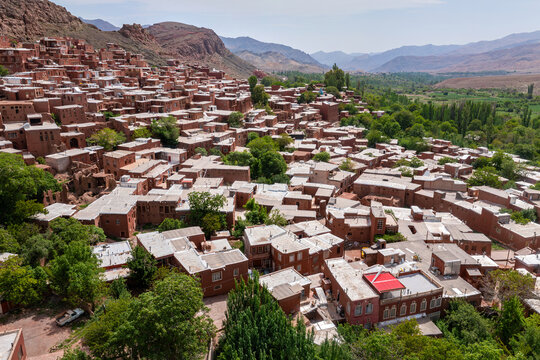 Aerial view of the red mud-brick houses cascading down the hillside, a tapestry of ochre against the rugged, mountainous backdrop, Abyaneh, Isfahan Province, Iran.