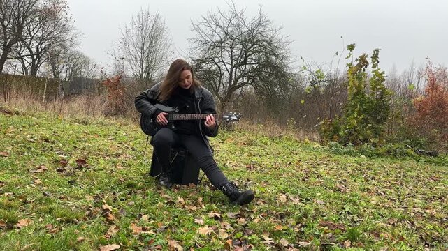 A female rock musician in a leather jacket plays an electric guitar with an amplifier in the garden in gloomy weather