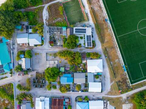 Aerial view of rooftops and lush greenery interspersed with solar panels, juxtaposed with the manicured sports field, Vashafaru, Haa Alif, Malediven.