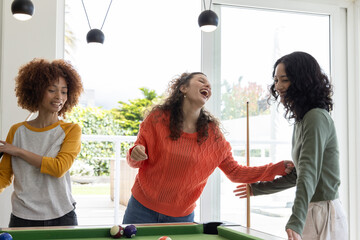 Diverse female friends laughing, playing at green pool table in game room with cue stick, balls
