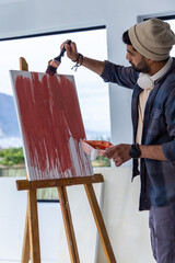 Male artist painting tall canvas on wood easel at studio wearing beanie denim shirt holding brush