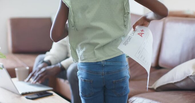 Student raising school report card showing letter A+ near parent using laptop, seeking parent pride