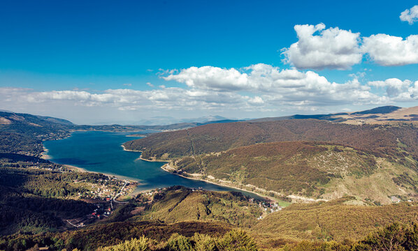 View of the tranquil lake reflecting the azure sky, embraced by rolling hills of lush forest under a blanket of fluffy clouds, Mavrove, North Macedonia.