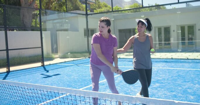 Two women practicing paddle and ball drills on fenced blue court, gray tank guiding purple player