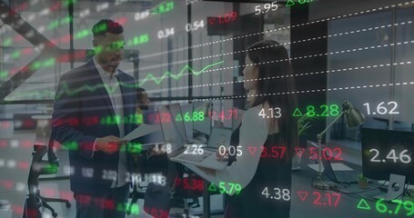 Reviewing papers, coworkers in blazer and vest holding laptop with financial overlay in open office