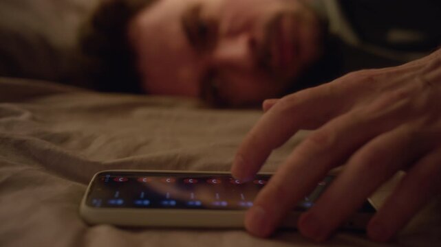 Rack focus shot of depressed adult man lying on bed checking smartphone for missed calls then burying face in pillows while struggling with social anxiety, copy space