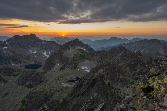 View of a radiant sun dips behind jagged peaks, casting a warm glow on the rugged terrain, while dark lakes mirror the sky's fiery hues, Vysoke Tatry, Presov Region, Slovakia.