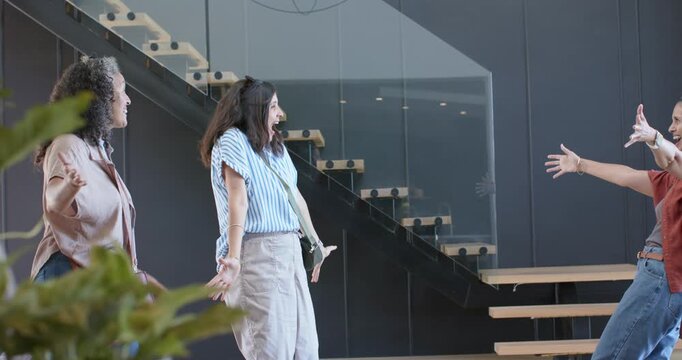 Diverse women reacting to 3rd woman arriving, embracing in lobby with suitcase, reuniting