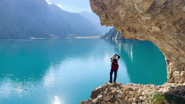 Horizontal video of a woman stands on a cliff by Tientang Hu lake, Xinjiang, China. The curved stone corner of the lakeside walkway. The sun was rising with yellow light reflected on the water.