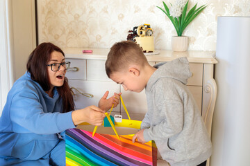 Obraz premium excited mother watching little boy opening colorful birthday gift bag during sixth birthday celebration at home with construction themed cake on background