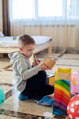 little boy sitting on floor opening small birthday gift box and holding candy during sixth birthday celebration at home with colorful gift bag and balloons