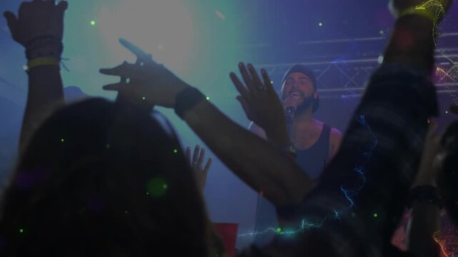 Raising backlit hands, crowd watching singer in tank top holding mic on stage, with particle swirls