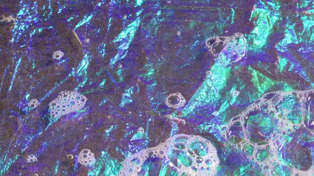 Irregular shapes ripple across iridescent surface. Color shifts from deep blue to emerald green. Foamy patches float on shimmering liquid. Abstract texture suggests underwater or mineral origin