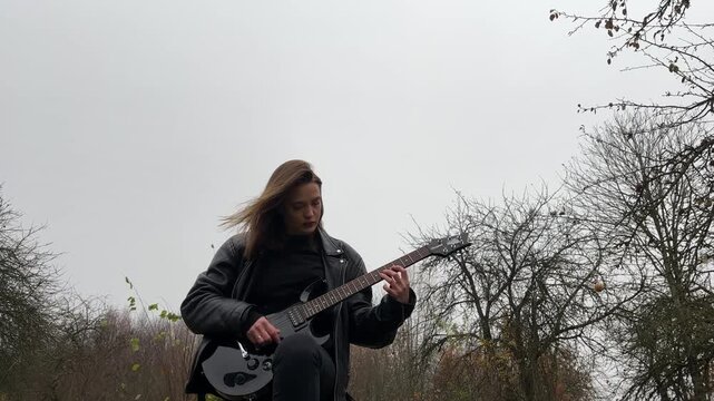 young female rock musician in a leather jacket plays an electric guitar outdoors