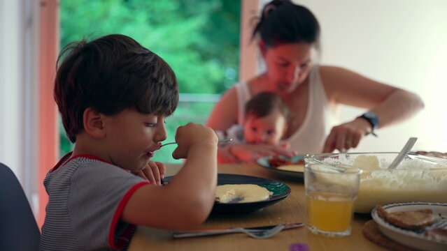 Family dining together, mother multitasking while older child eats independently, home-cooked meal, nurturing environment, parent balancing caregiving and mealtime