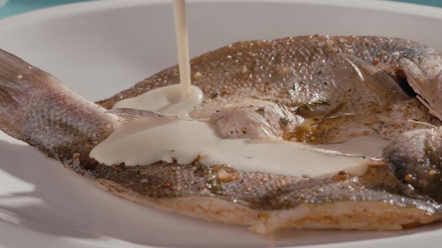 Pouring creamy sauce over whole flatfish served on white plate. Elegant gourmet seafood dish with restaurant style presentation and fine dining plating
