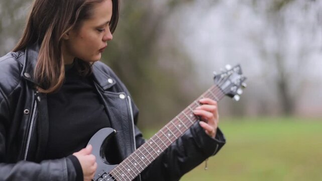 A female rock musician in a leather jacket plays an electric guitar outdoors in gloomy weather