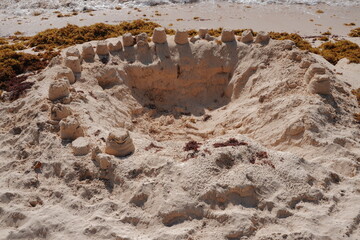 Wide high-angle shot of a large sandcastle structure with turrets on a tropical beach. Background features scattered sargassum seaweed and ocean waves under bright daylight. © 1975boomerang