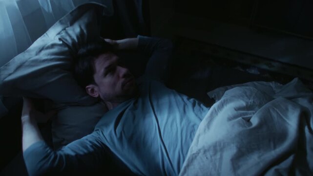 Middle aged man suffering from sleep deprivation feeling discomfort while lying in bed, tossing pillow up to get to sleep at late night