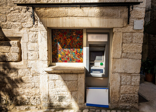 Trogir, Croatia &ndash; March 7, 2026: ATM cash machine installed in a historic stone wall next to a decorative stained glass window in the old town of Trogir, Dalmatia, Croatia.