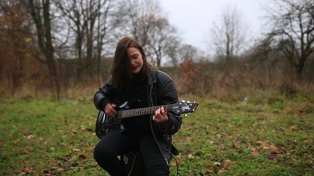 young female rock musician in a leather jacket plays an electric guitar outdoors