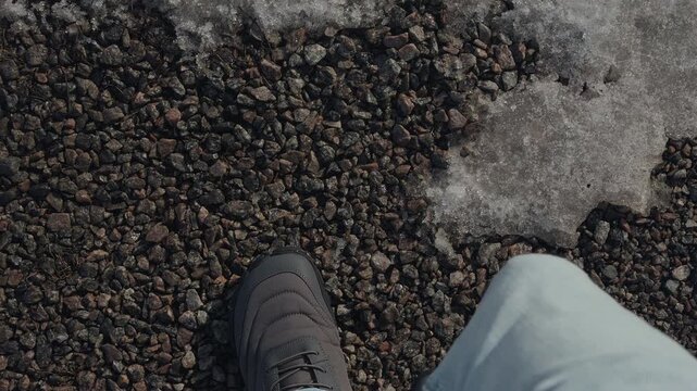 A top-down view of a person wearing grey winter boots and light blue jeans, walking on dark gravel next to patches of melting snow. Close up.