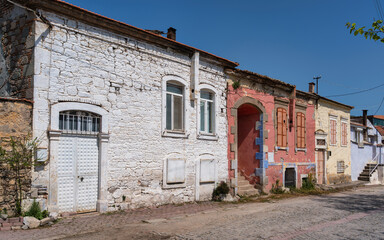 Old houses line a cobblestone street in a village. Some houses have colorful walls, and many show signs of age. The sun shines brightly above, highlighting the simple structures.