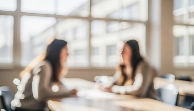 Two people engaged in a discussion at a table in a bright room with large windows. The image has a soft focus, creating a warm and inviting atmosphere.