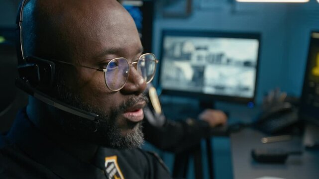 Side view close up shot of African American man working as dispatcher at police command center and answering calls via headset while sitting at desk and looking at computer monitors during night shift