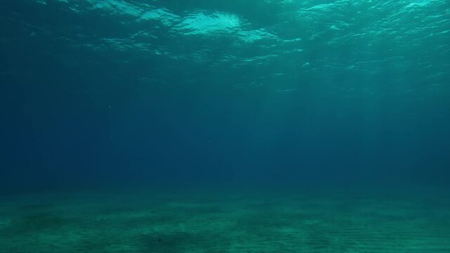 Slow motion forward of underwater sun light shine under deep water with ripples on waves surface, God rays penetrate through waves on water surface and fall in glare on sandy sea bottom