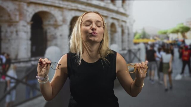 Woman blonde testing two pairs of eyeglasses, holding frames by each hand, lips puckered and comparing fit while standing before an ancient stone building and busy street; playful choice.