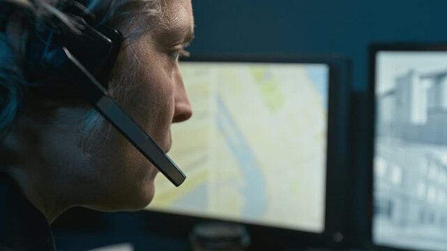 Over shoulder close up shot of mature woman working as dispatcher at police station and answering calls via headset while watching computer monitors with CCTV feed and city maps