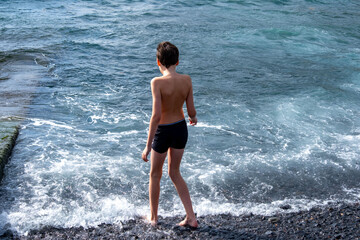 Fototapeta premium Teen boy playing in ocean waves on black volcanic sand beach, joyful summer moment, natural fun, healthy childhood, barefoot freedom