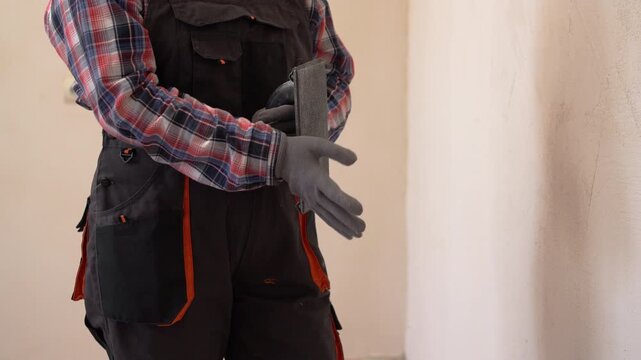 repairwoman in work overalls preparing for home renovation wearing gloves and holding drywall sanding tool for smoothing putty on walls indoors, construction work before finishing stage