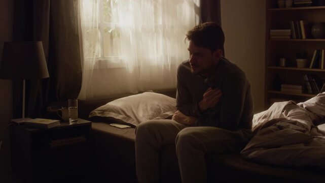 Adult man getting out of bed feeling tightness in chest then drinking water to soothe panic attack in dimly lit bedroom