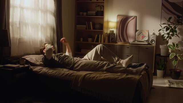 Wide angle shot of middle aged man feeling morning fatigue when waking up then curling up on bed with pillow in sunlit bedroom