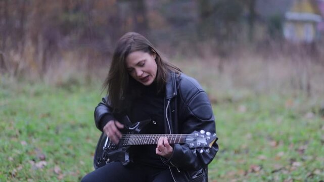 A female rock musician in a leather jacket plays an electric guitar outdoors in gloomy weather