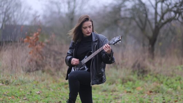 A female rock musician in a leather jacket plays an electric guitar outdoors in gloomy weather