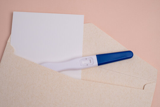 Blue tip midstream pregnancy test and blank invitation card inside beige textured envelope over pink background. Pregnancy announcement concept. 