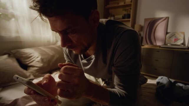 Handheld shot of stressed middle aged man scrolling news feed on smartphone then putting device away feeling drained and desperate while sitting on bed in dimly lit room