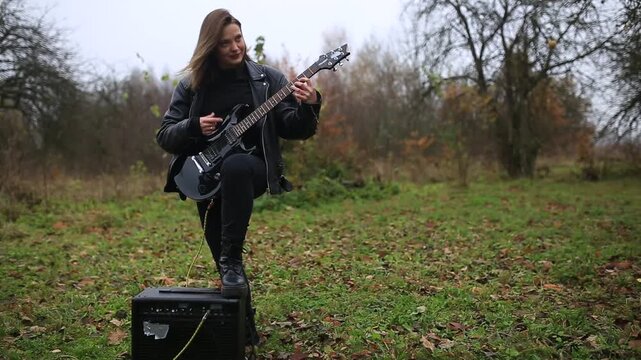 A female rock musician in a leather jacket plays an electric guitar outdoors in gloomy weather