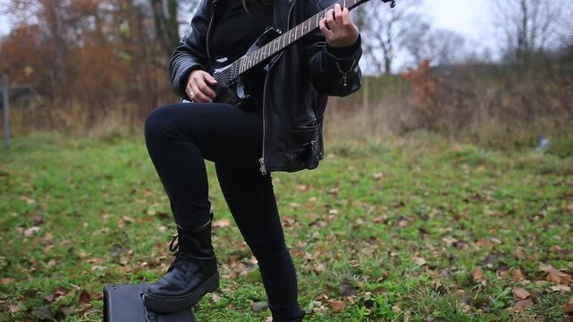 A female rock musician in a leather jacket plays an electric guitar outdoors in gloomy weather